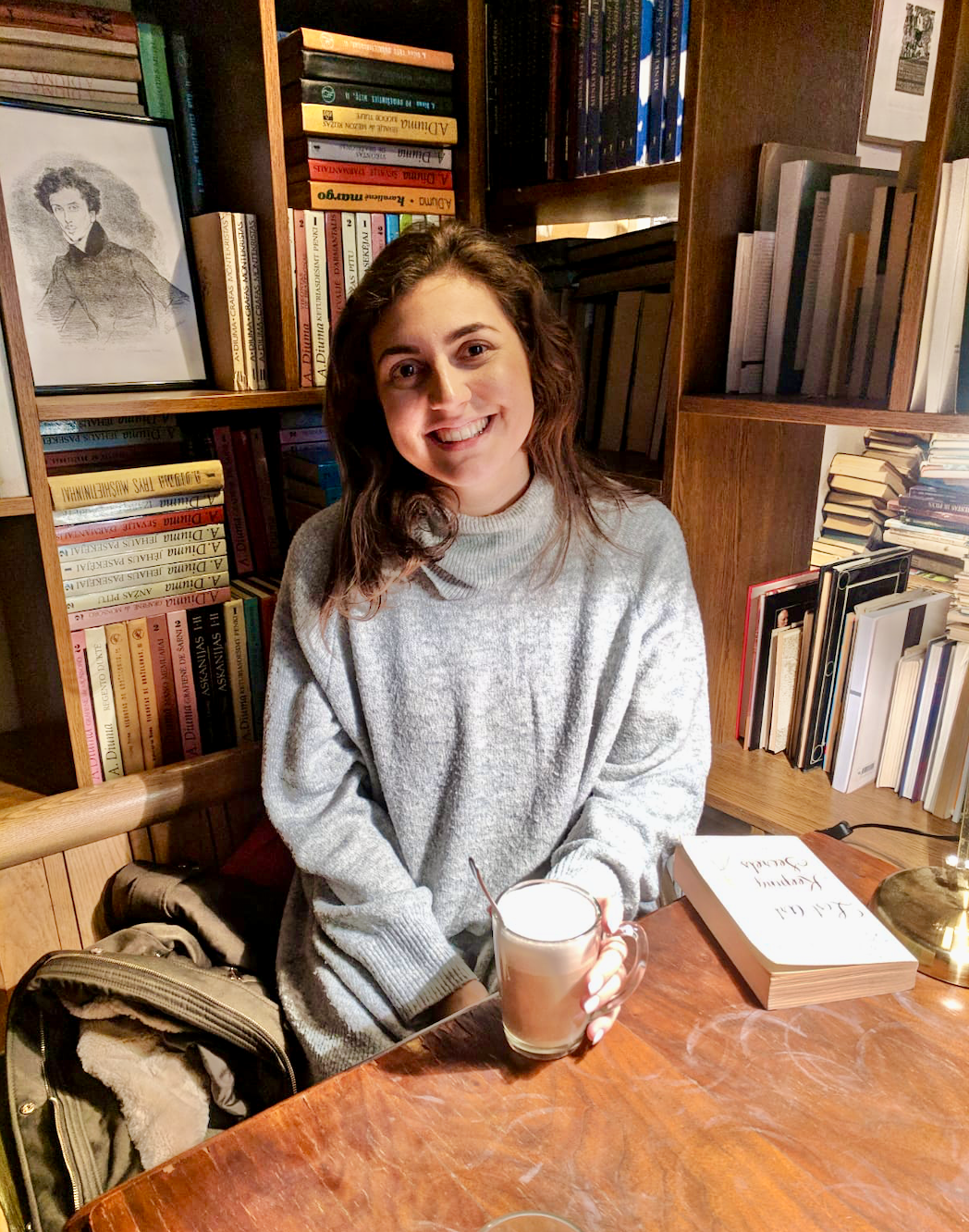 Best Restaurants in Vilnius: Nell sat in a cafe surrounded by shelves of books with a coffee in her hand. She's looking at the camera and smiling