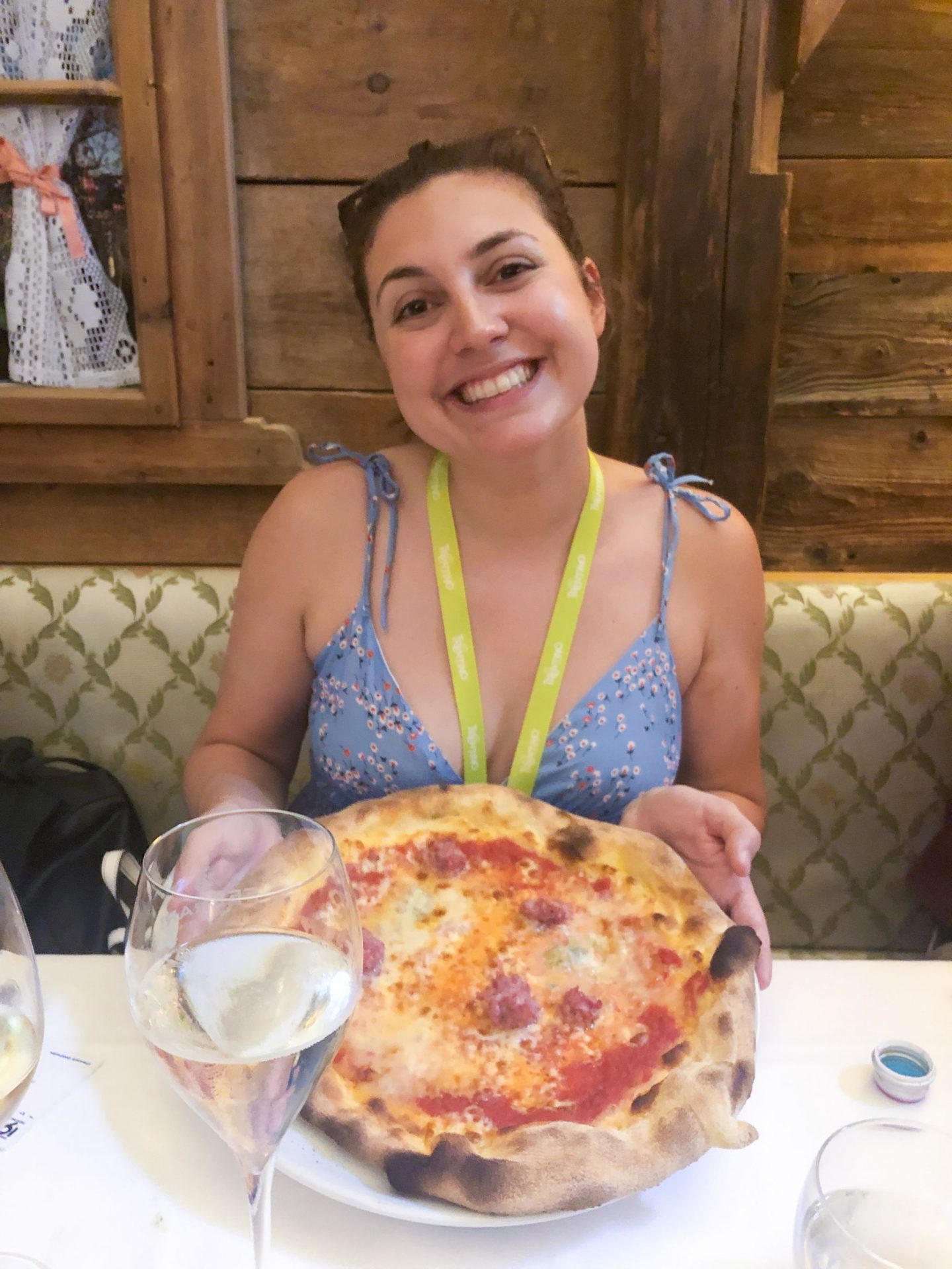 Girl smiling holding up a whole pizza with a glass of wine in front of her, eating and drinking in Trento, Trentino