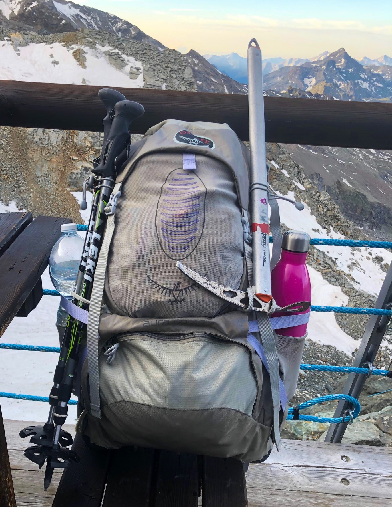 Osprey Aura Anti Gravity Rucksack with mountains in the background. Attached to the rucksack is an ice axe and walking poles, and a pink water bottle is in the side pocket.