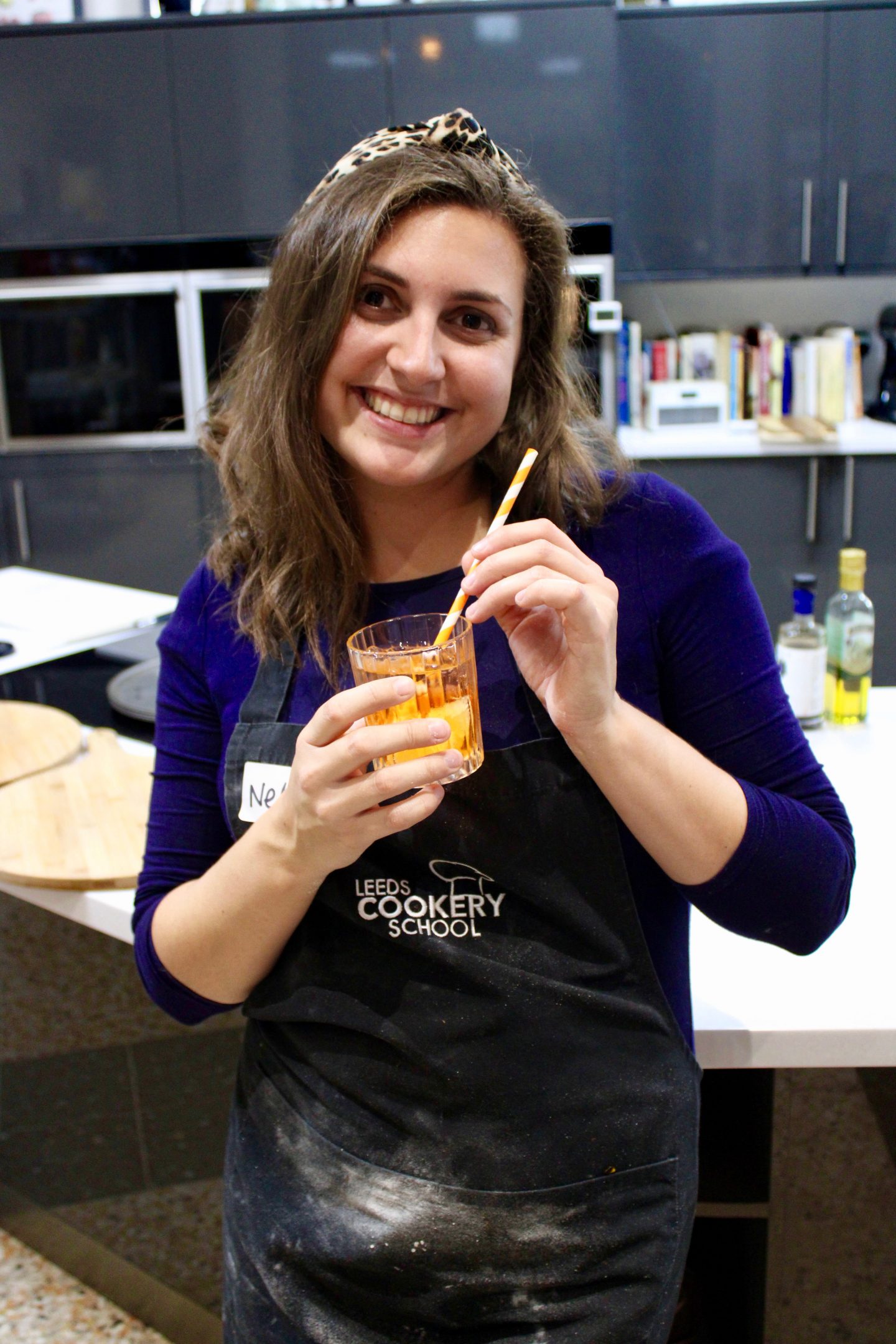 pizza making class leeds cookery school: Nell wearing blue dress and leopard print headband smiling at the camera whilst holding a glass of Aperol Spritz. In the background a kitchen scene is just out of focus.