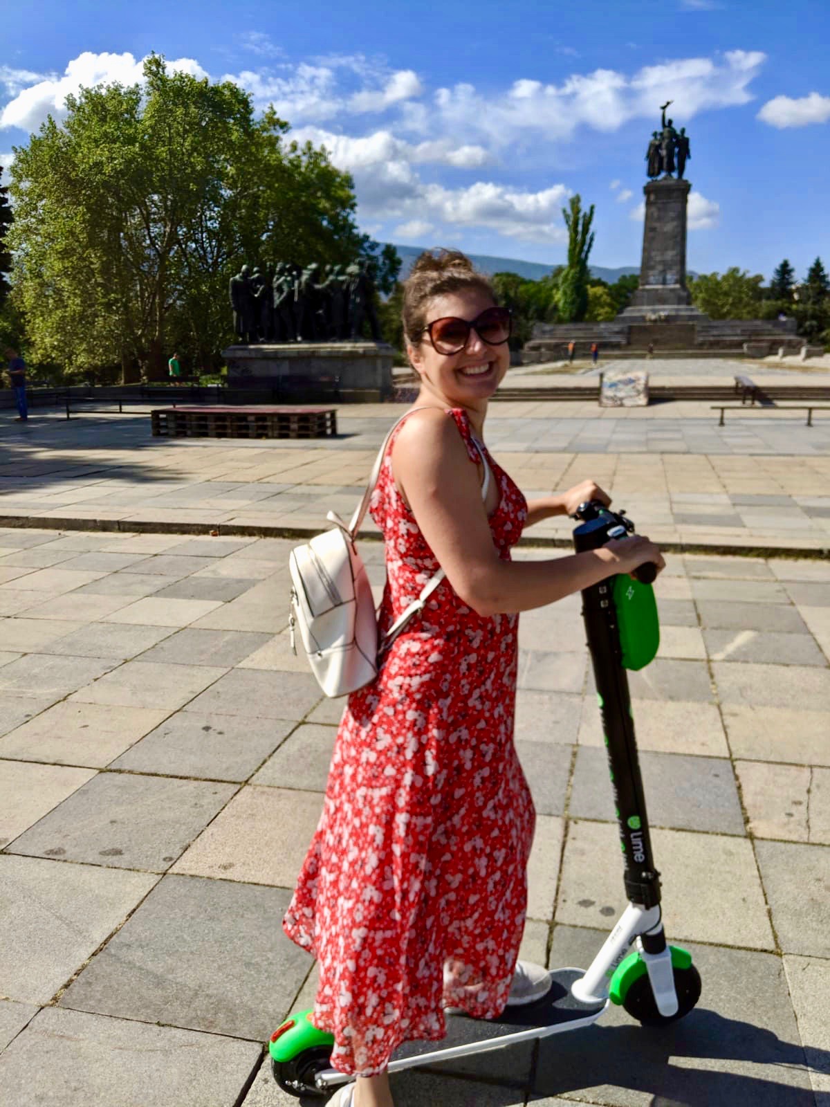 Nell in red floral dress on an electric scooter, in a park in Sofia enjoying her city break. There is a statue behind her and the sky is blue, she has one leg on the scooter and is turned to smile at the camera