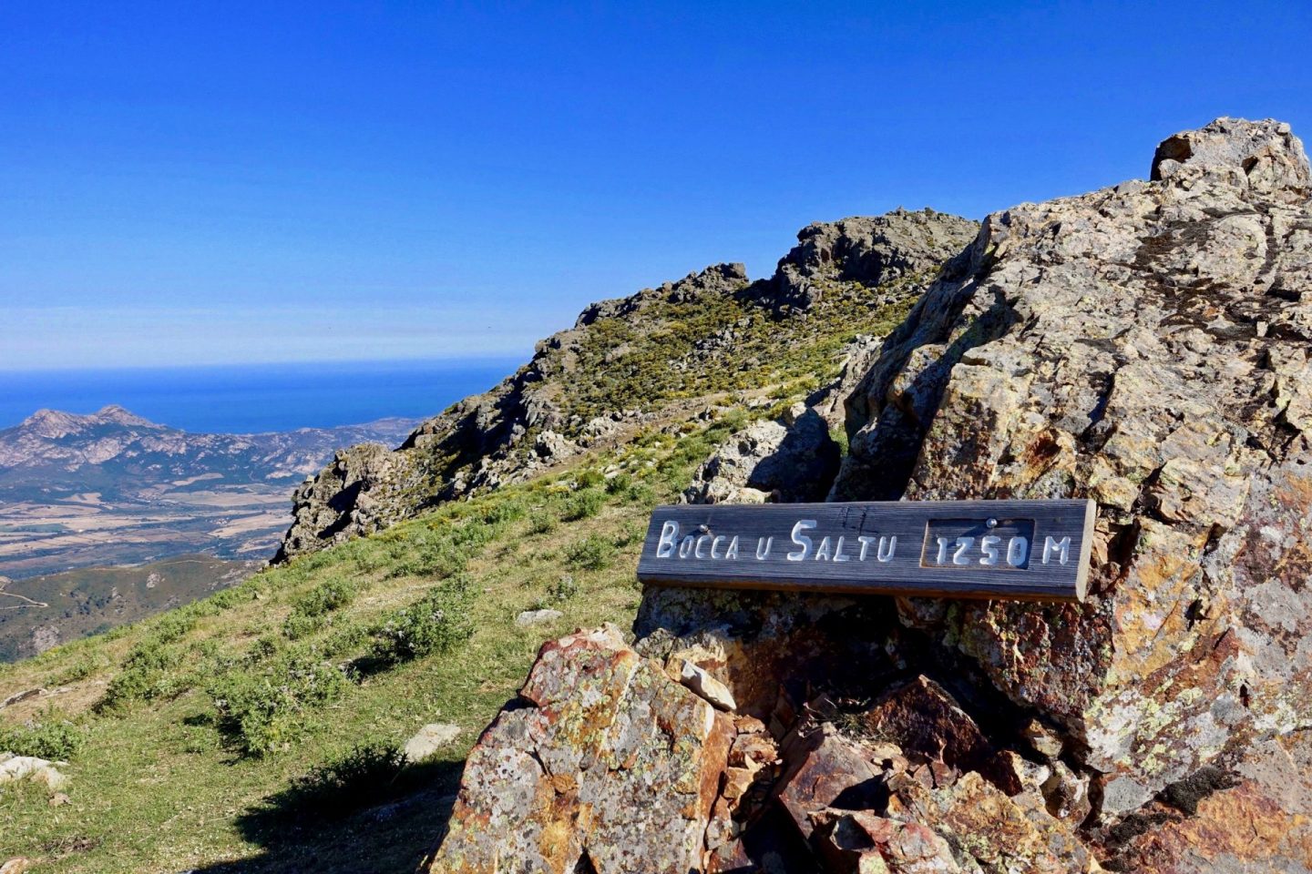 A wooden sign saying Bocca u Saltu 1250 m, bolted to rock. The sea can be seen in the distance behind.