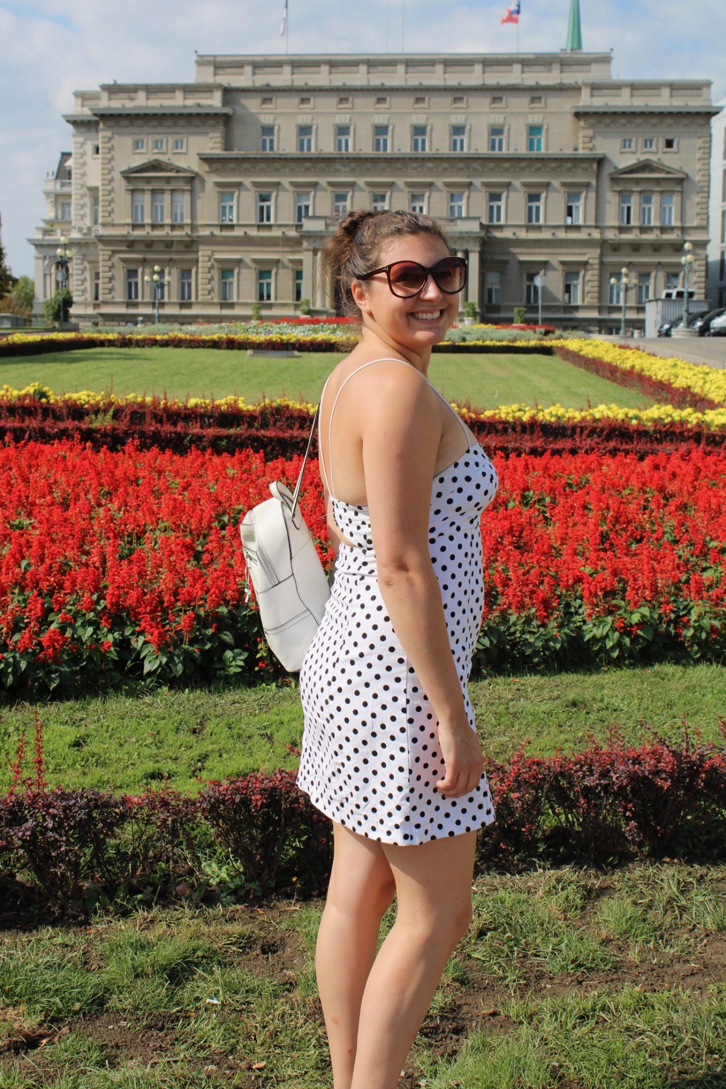 cheap european city break - Nell wearing a white polkadot dress, stood in front of flowers and an impressive building whilst on holiday in Belgrade