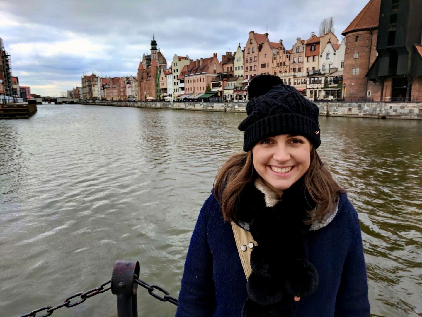 cheap european city break - Nell in Gdansk smiling at the camera and wearing a black hat. Behind her is water and then a beautiful row of buildings