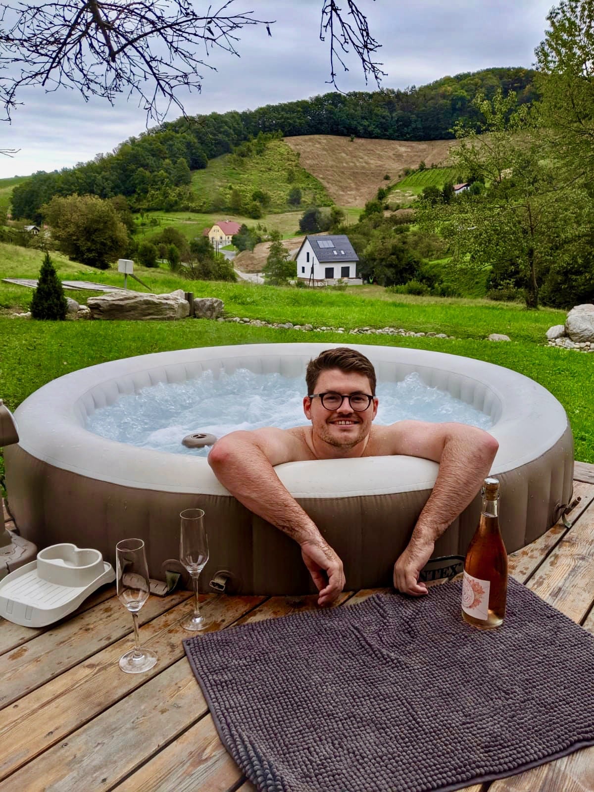 Glamping in Slovenia: Man in hot tub smiling at the camera, with vineyards in the background and a bottle of sparkling wine in front of him